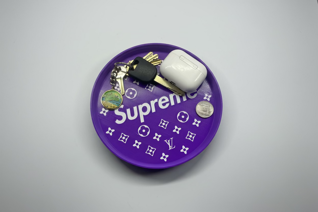 Purple round tray with 'Supreme' branding, keys, and a white air pod case on a gray surface.